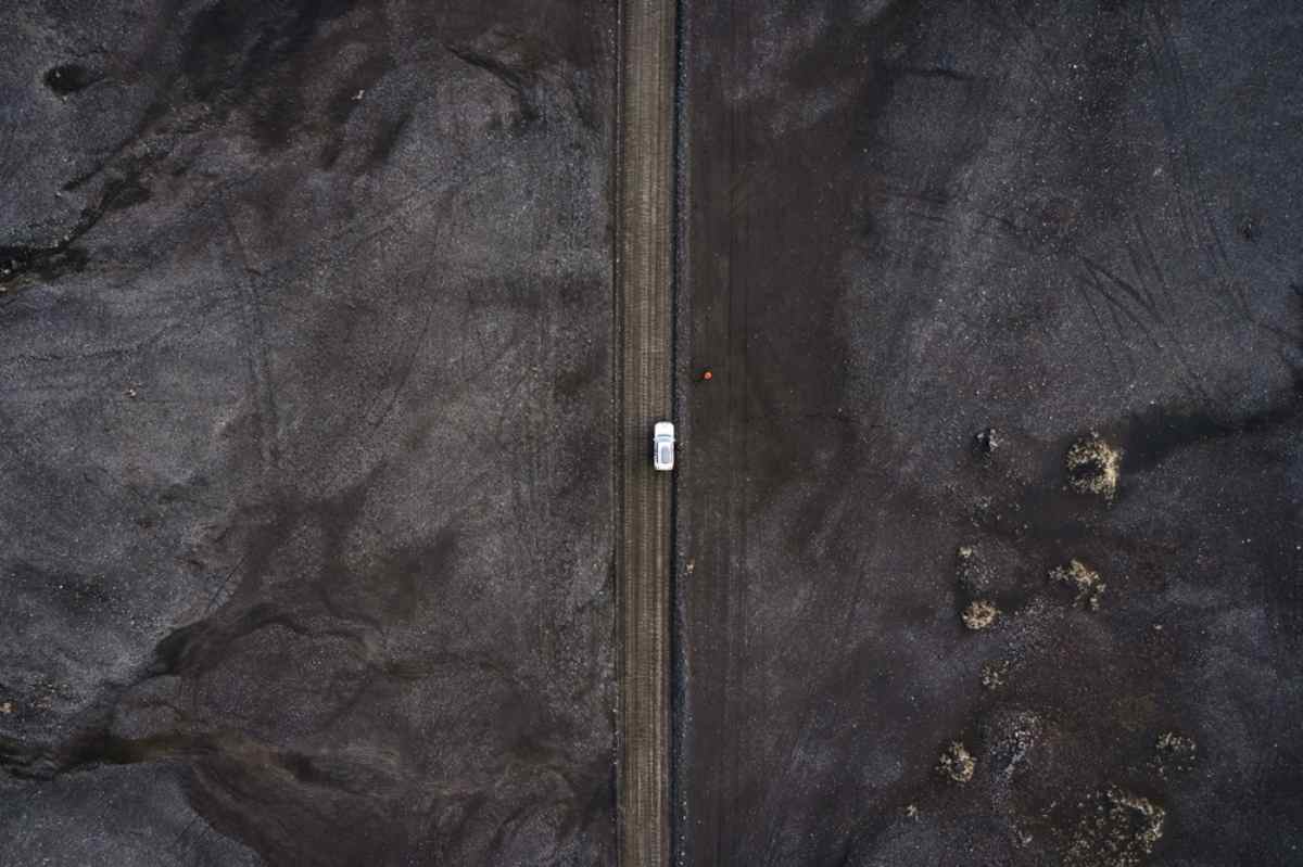 Vista aerea di un veicolo bianco che attraversa quella che sembra una strada islandese non asfaltata piena di sabbia nera e cenere.