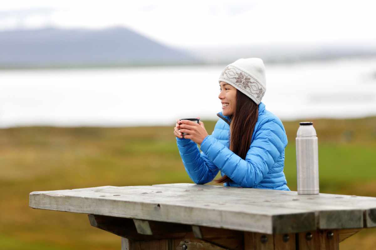 Persona sorridente con piumino blu e berretto di lana che sorseggia una bevanda calda a un tavolo da picnic in Islanda, con un thermos e colline nebbi