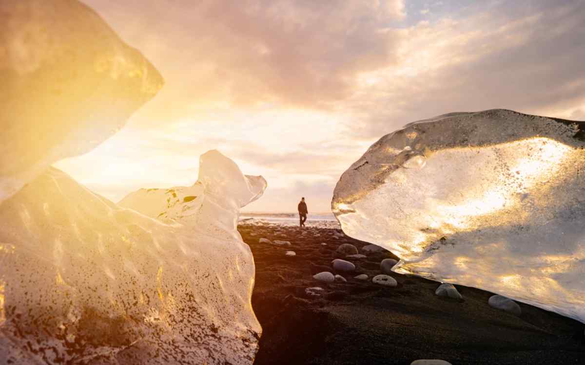 Iceland's Diamond Beach Traveler walking along Iceland’s Diamond Beach at sunrise, framed by glowing ice chunks scattered across the black sand.