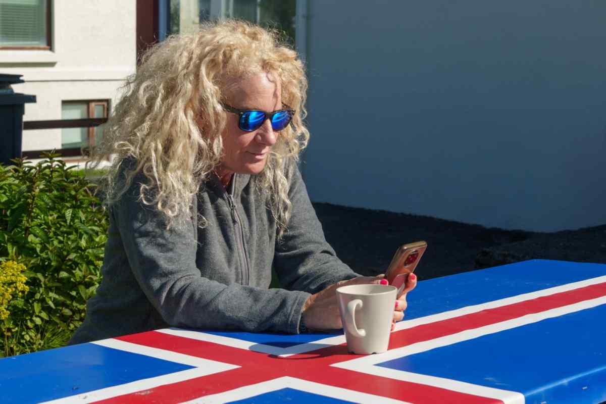 Warm temperatures in Iceland Woman relaxing with a coffee at a table painted with the Icelandic flag on a sunny summer day, checking her phone outdoors.