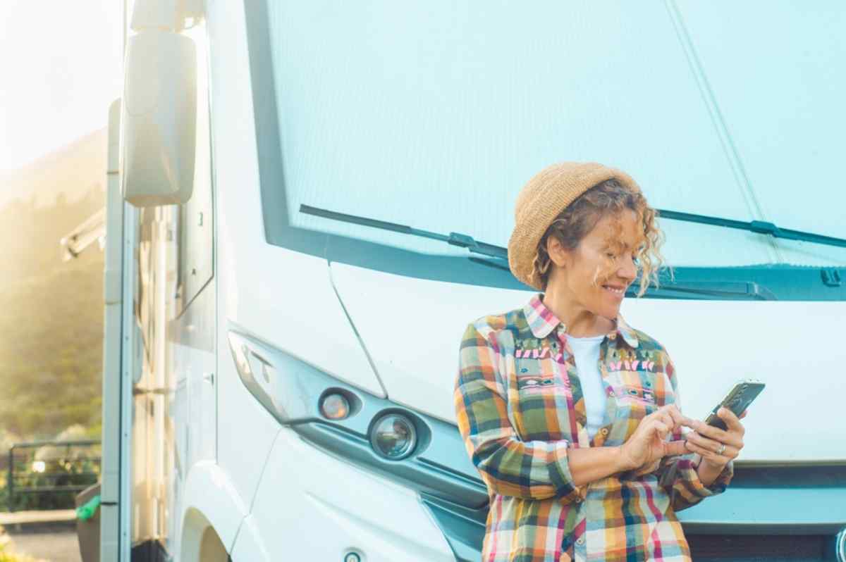 Tips for camping in Iceland Smiling traveler checking her phone beside a parked campervan at sunset, planning her next stop during a relaxed summer road trip.