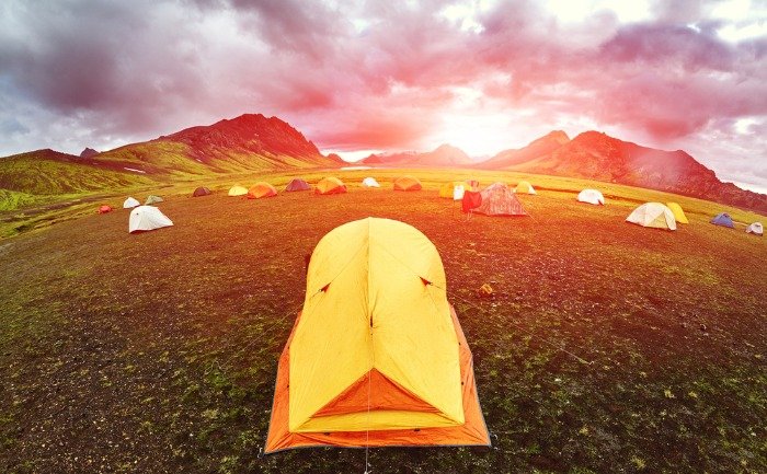 Tienda de campaña amarilla en primer plano en un gran camping al aire libre, con más tiendas repartidas por el terreno y montañas al fondo bajo un cie