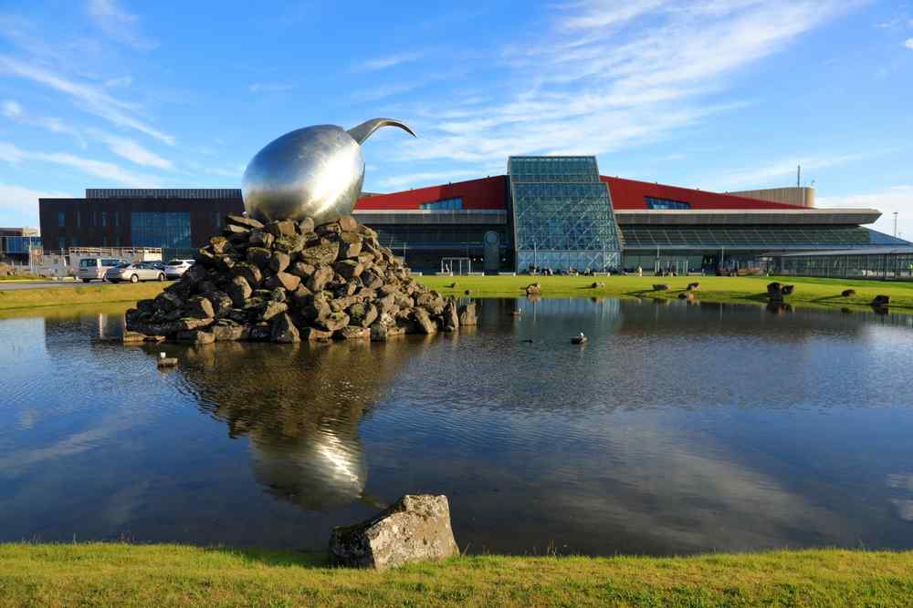 Esterno dell’aeroporto internazionale di Keflavík con uno specchio d’acqua riflettente e una grande scultura metallica a forma di uovo davanti al term