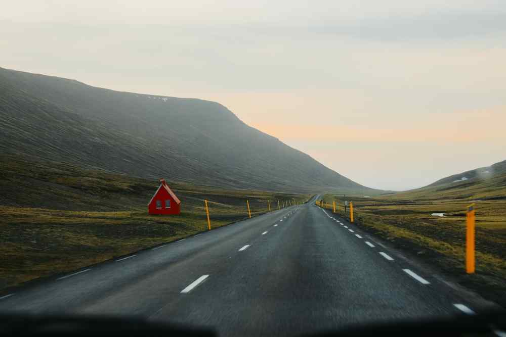 Empty paved road stretching through a remote Icelandic valley, with mountains, roadside markers and a small red house.