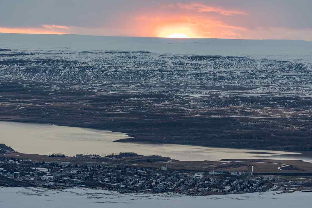 Iceland's Egilsstaðir Snow-covered view of Egilsstaðir in East Iceland beside Lagarfljót lake, with the sun setting behind the surrounding mountains.
