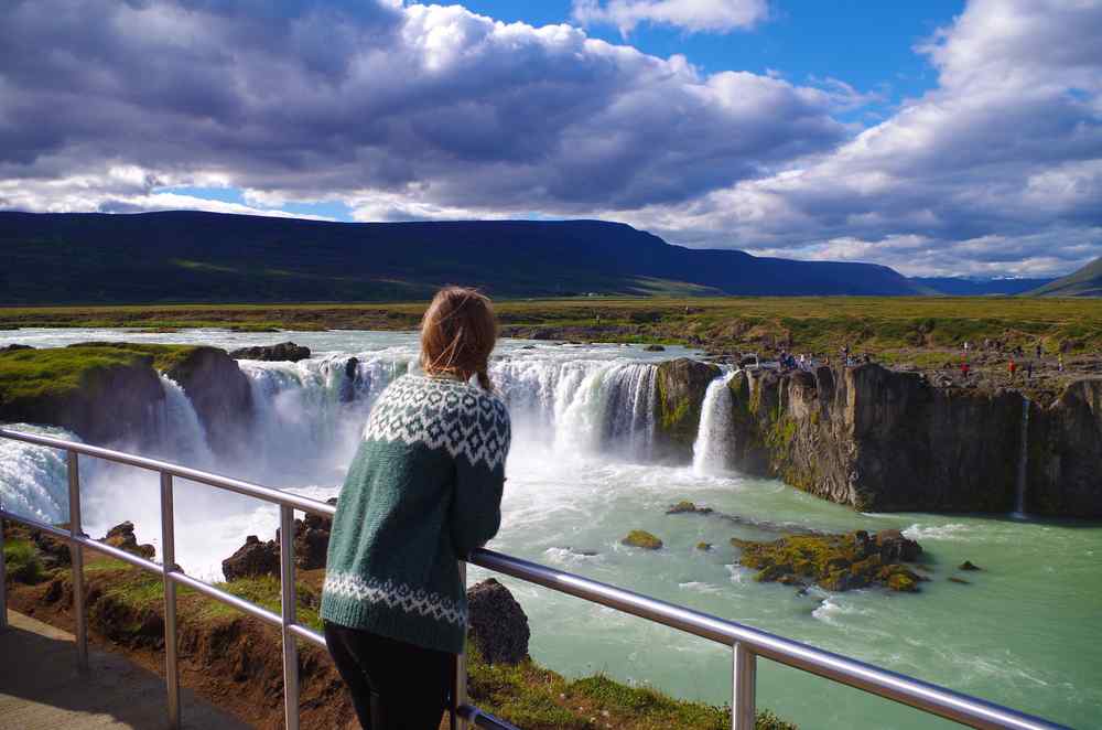 Iceland's Godafoss waterfall Traveler wearing an Icelandic sweater looking over the powerful Goðafoss waterfall beneath dramatic clouds in northern Iceland.