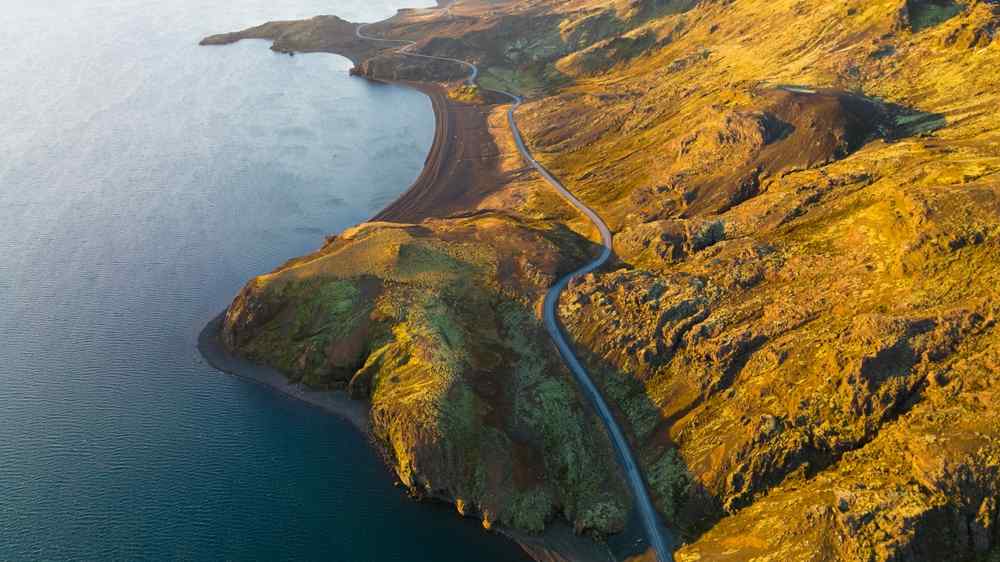 Iceland's Golden Circle Aerial view of a winding road following Iceland’s rugged coastline, surrounded by volcanic hills illuminated by golden sunlight.