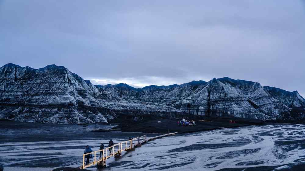 Ice caving in Iceland Visitors walking across a boardwalk through a dramatic volcanic landscape surrounded by striped mountains in Iceland.