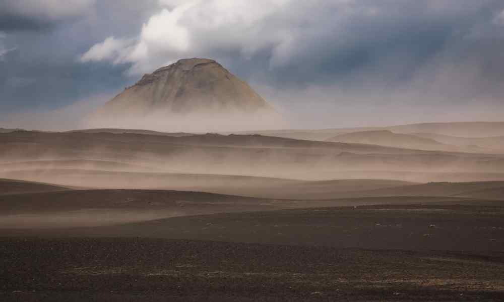 The importance of sand and ash protection in Iceland Strong wind blowing volcanic sand and ash across a barren Icelandic landscape with a mountain partly obscured in the distance.