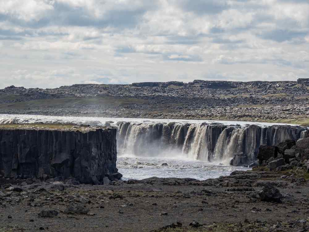 Iceland's Selfoss Wide Icelandic waterfall flowing over dark basalt cliffs in a barren volcanic landscape beneath a cloudy sky.