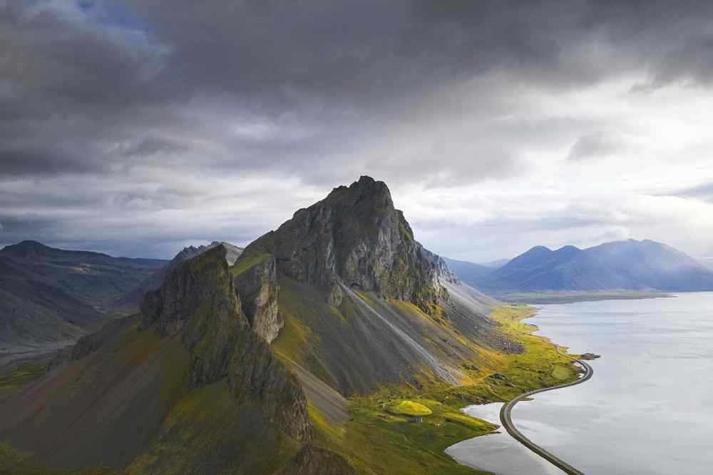 South Coast in Iceland Dramatic mountain ridge rising beside a winding coastal road and calm sea under stormy skies in South Iceland.