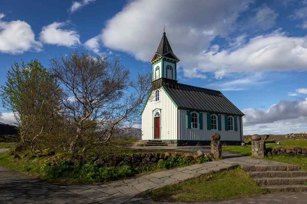Iceland's Thingvellir National Park Historic white and green church at Þingvellir National Park surrounded by trees and lava-stone walls under a blue sky.