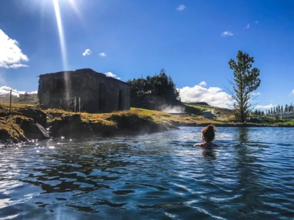 Person enjoying a geothermal hot spring in rural Iceland, with a rustic building and lush greenery under a bright blue sky and sunlight