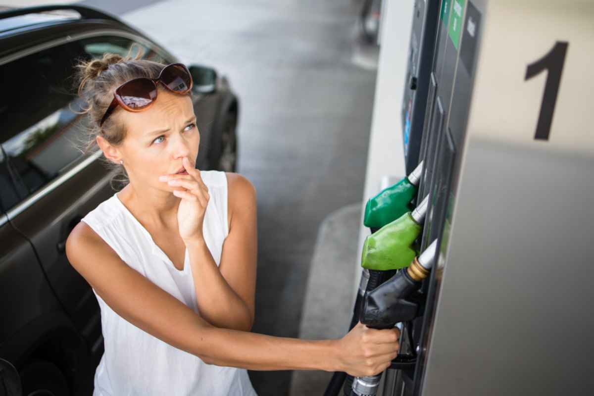 A blonde woman with her hair tied back looks worriedly at the price of gas while holding a pump at a gas station.