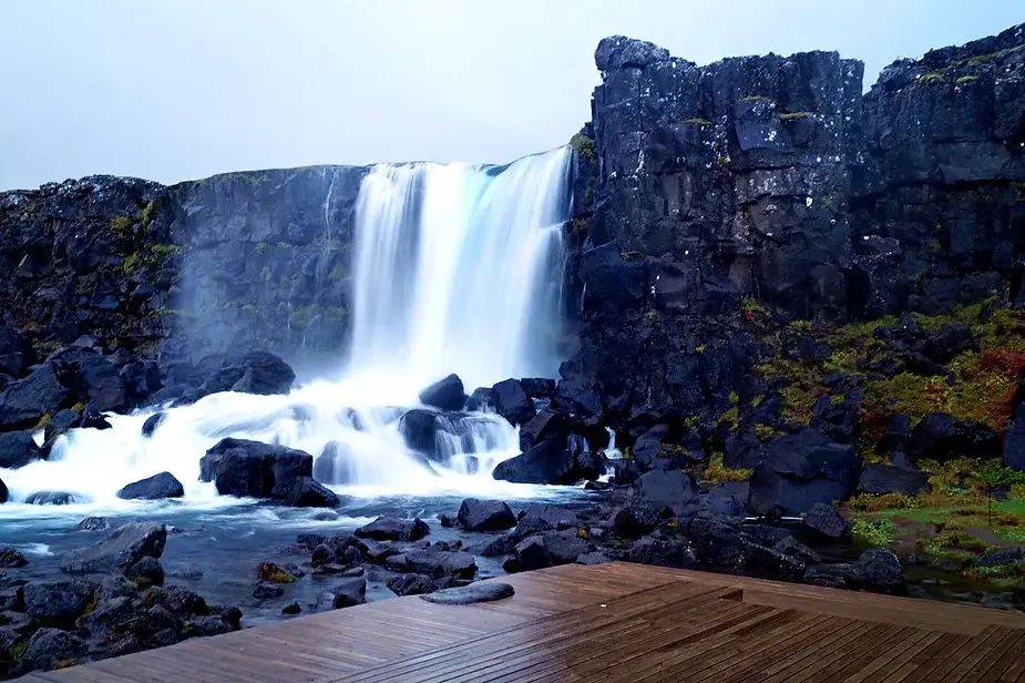A wooden boardwalk leading to Öxarárfoss Waterfall, flowing against the backdrop of volcanic cliffs in Thingvellir National Park, Iceland. The area is