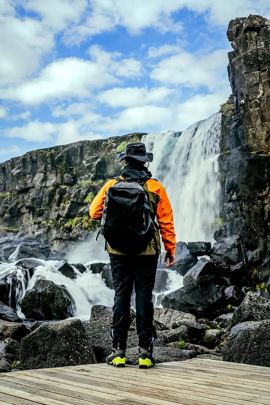 A person wearing outdoor gear and standing on a wooden platform, gazing at Öxarárfoss Waterfall in Thingvellir National Park, Iceland. The waterfall cascades down black volcanic rocks under a partly cloudy sky.