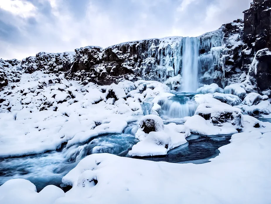 A snow-covered landscape surrounding Öxarárfoss Waterfall, with ice forming on the cliffs and rocks. The waterfall flows despite the thick winter frost, creating a stunning winter scene in Thingvellir National Park, Iceland.