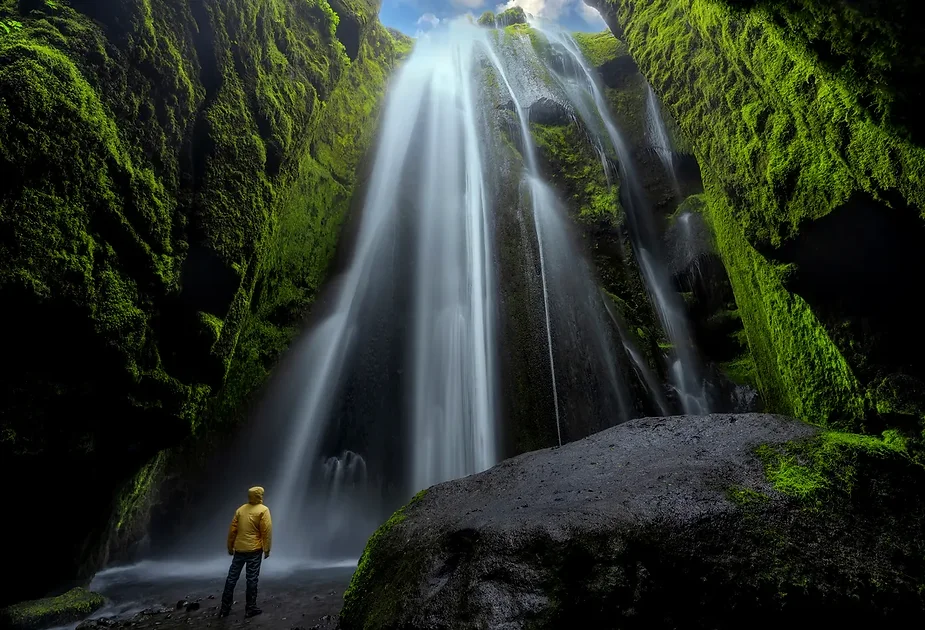 Majestic Gljúfrabúi waterfall cascading through moss-covered cliffs in Iceland, with a person in a yellow jacket admiring the natural beauty.