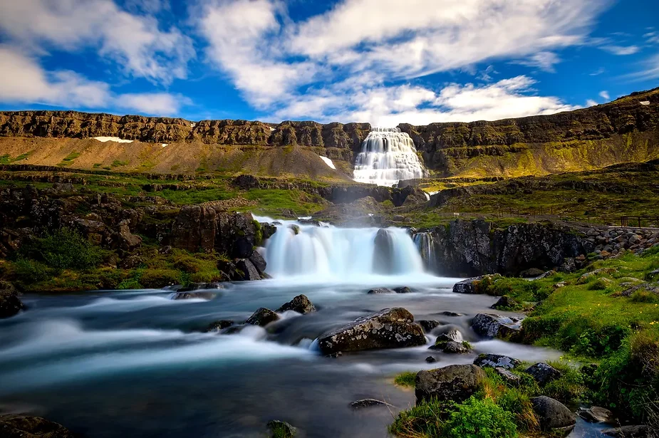 Dynjandi waterfall in Iceland cascading down multiple tiers of cliffs into a calm river, surrounded by lush greenery under a bright blue sky.