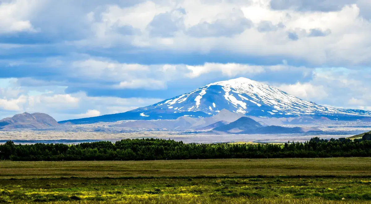The snow-capped peak of Hekla Volcano rises in the distance, with its slopes framed by lush green plains and a vibrant blue sky dotted with clouds.