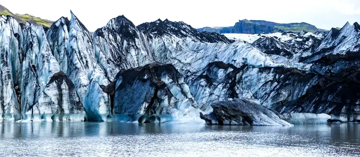 A glacier with jagged, black-and-white streaked ice formations rises from the water, set against a backdrop of green mountains and distant peaks in Iceland.