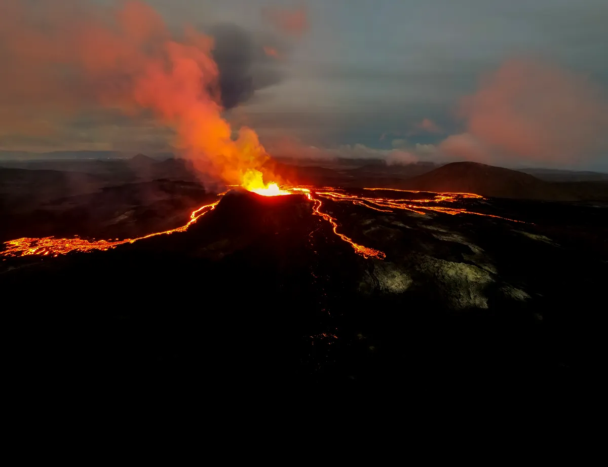 A stunning aerial view of a volcanic eruption at night, with bright red lava flows carving their way through dark volcanic rock.