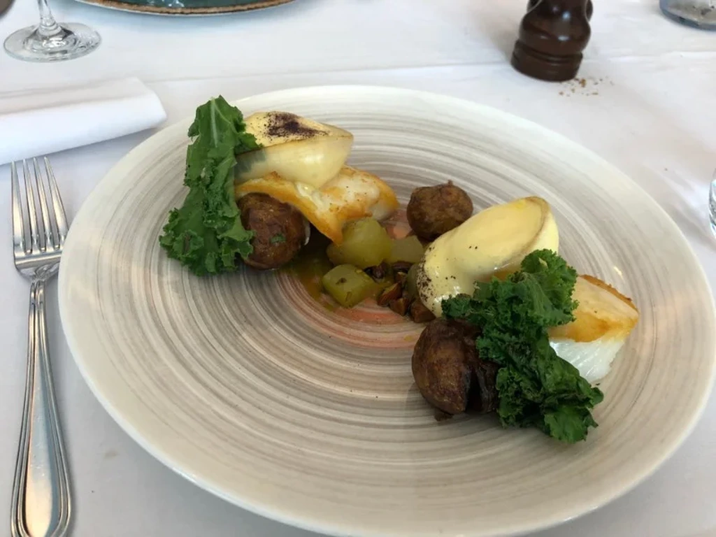 A gourmet dish served on a beautifully designed plate. The meal includes a portion of seared fish with melted cheese on top, accompanied by roasted baby potatoes, kale, and a selection of pickled vegetables. The elegant presentation is complemented by a white tablecloth, a neatly placed fork, and a glass of water visible in the background.