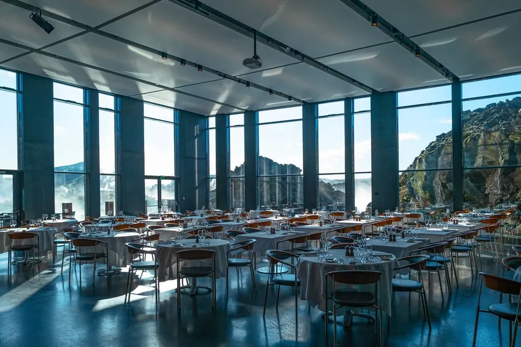 A modern restaurant with large floor-to-ceiling windows offering a stunning view of the volcanic landscape outside. The elegant setup includes round tables covered in white tablecloths, surrounded by neatly arranged wooden chairs, with crystal wine glasses and silverware placed perfectly on each table. The natural light flooding the room enhances the serene and upscale atmosphere.