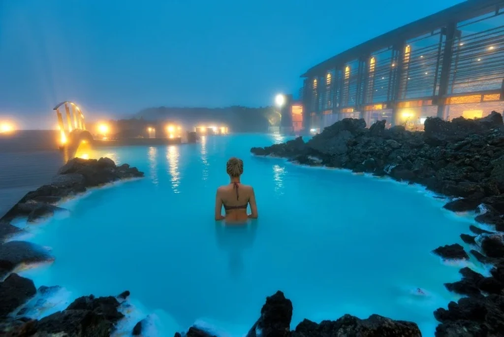 A serene view of the Blue Lagoon in Iceland, with a woman relaxing in the milky-blue geothermal waters. The warm light from the surrounding buildings and the misty atmosphere create a calm and tranquil ambiance, as the natural volcanic rock formations border the lagoon.