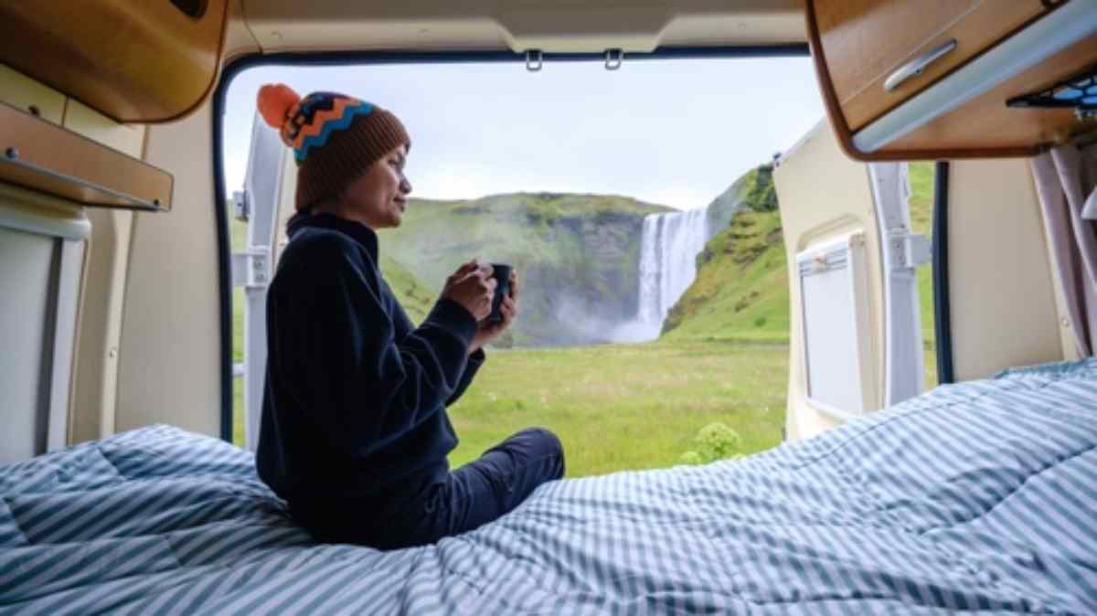 Person sitzt in einem Campervan mit einer Tasse Kaffee und blickt auf den Wasserfall Skógafoss in Island.