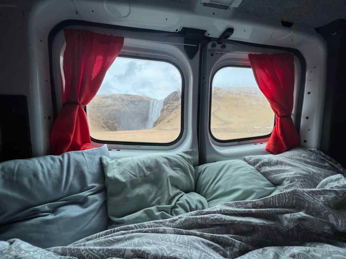 Blick auf einen Wasserfall durch das Heckfenster eines Campervans mit Kissen und Decken.