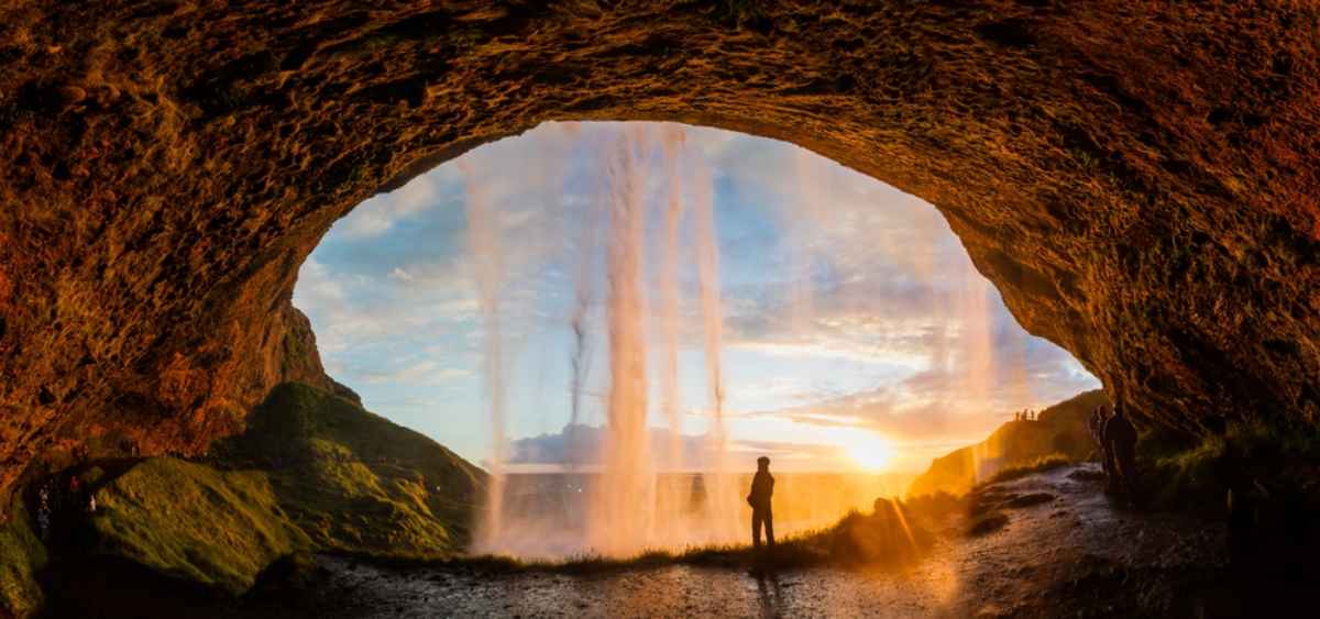 Reisende Person, die in einer H&ouml;hle hinter einem Wasserfall im Sonnenuntergang an der S&uuml;dk&uuml;ste Islands steht.