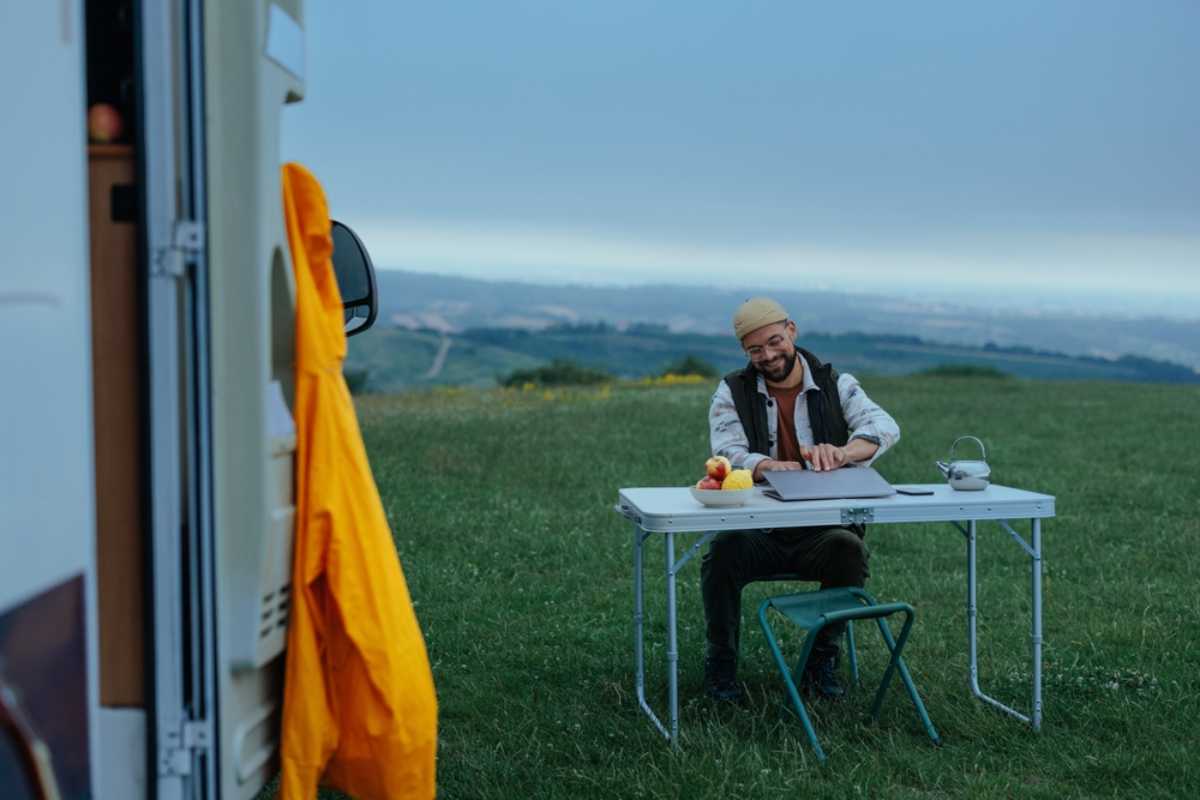 Reisender sitzt an einem Klapptisch neben einem Wohnmobil auf einer Wiese und arbeitet drau&szlig;en mit offenem Laptop