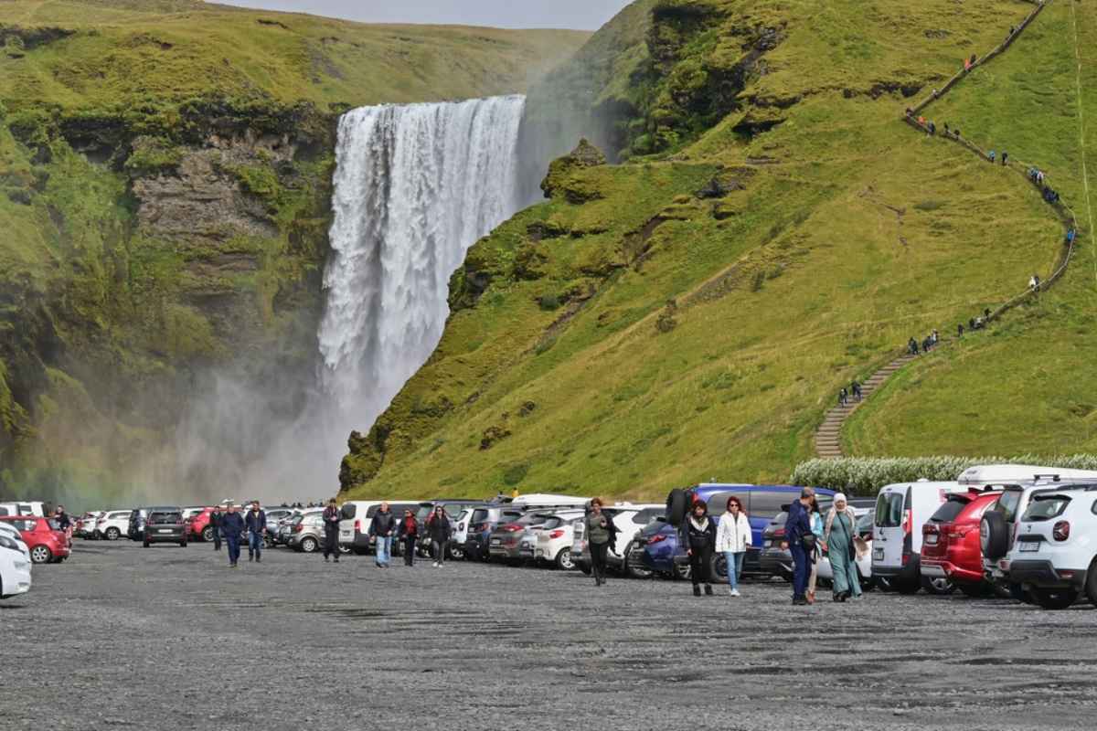Voller Parkplatz neben dem Wasserfall Skogafoss, w&auml;hrend Besucher zwischen geparkten Autos hindurchlaufen