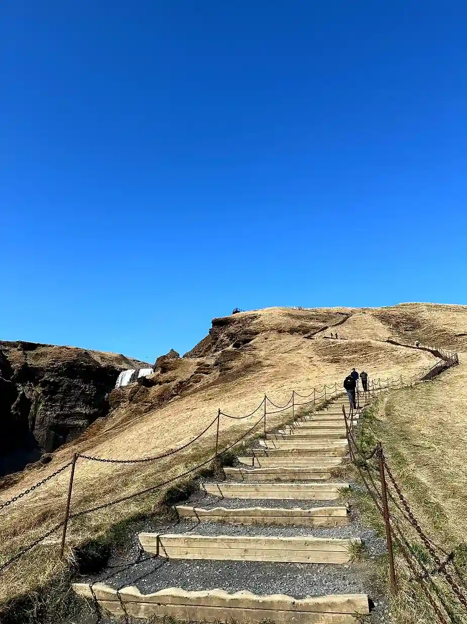 Touristen, die die Holztreppen hinauf zur Spitze des Skógafoss-Wasserfalls in Island gehen, mit einem klaren blauen Himmel und trockenen, grasbewachsenen Hügeln im Hintergrund