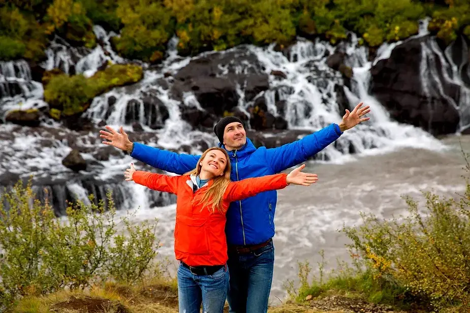 Happy couple in colorful jackets posing with outstretched arms in front of Hraunfossar waterfall in Iceland, surrounded by lush green foliage