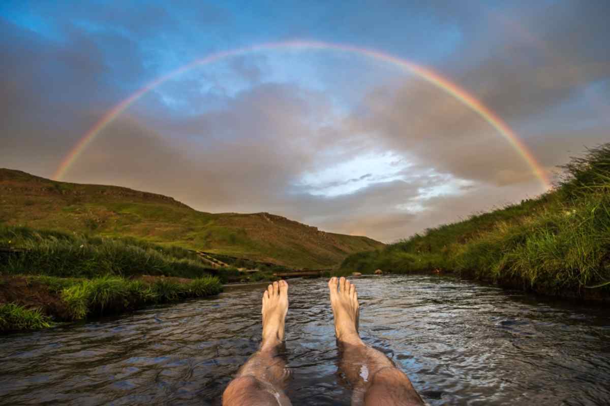 Una persona, descalza, se baña en un río islandés mientras se forma un arcoíris en el cielo.