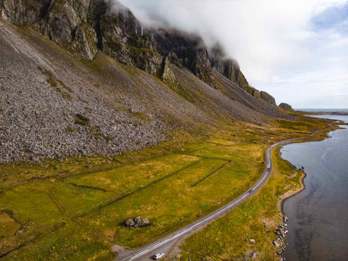 La Ring Road de Islandia Vista aérea de una carretera costera bordeando una ladera rocosa y empinada con nubes bajas, campos verdes y un fiordo en calma a lo largo de la Ring Road de Islandia.