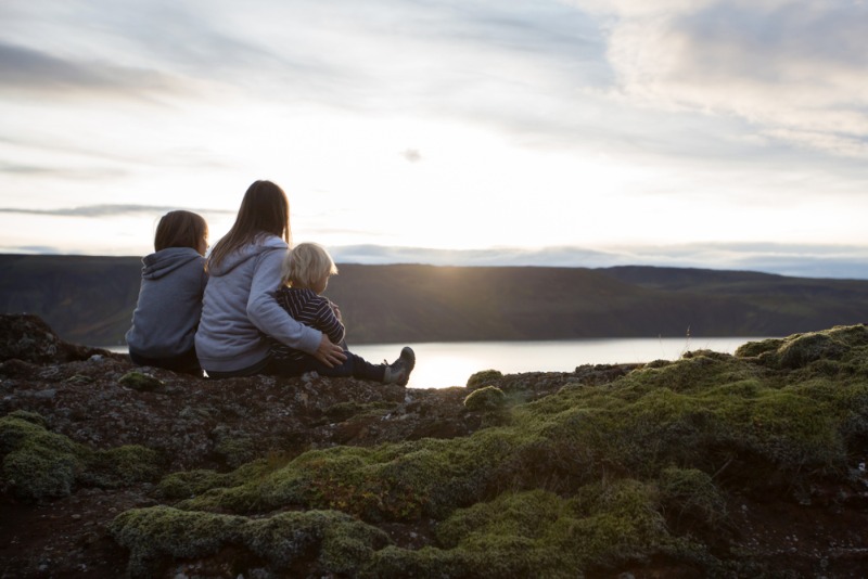 Madre con dos hijos sentados sobre rocas de lava cubiertas de musgo, mirando un lago islandés tranquilo y montañas al atardecer durante un viaje en ca
