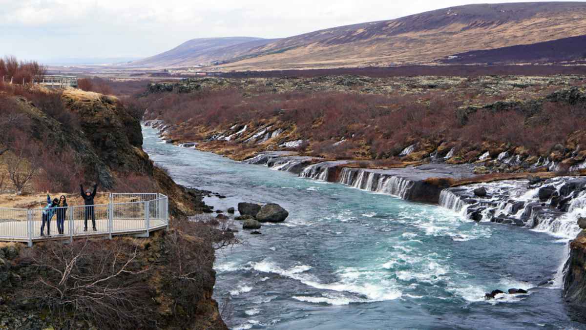 Familia saludando desde un mirador sobre un r&iacute;o turquesa y peque&ntilde;as cascadas en Islandia.