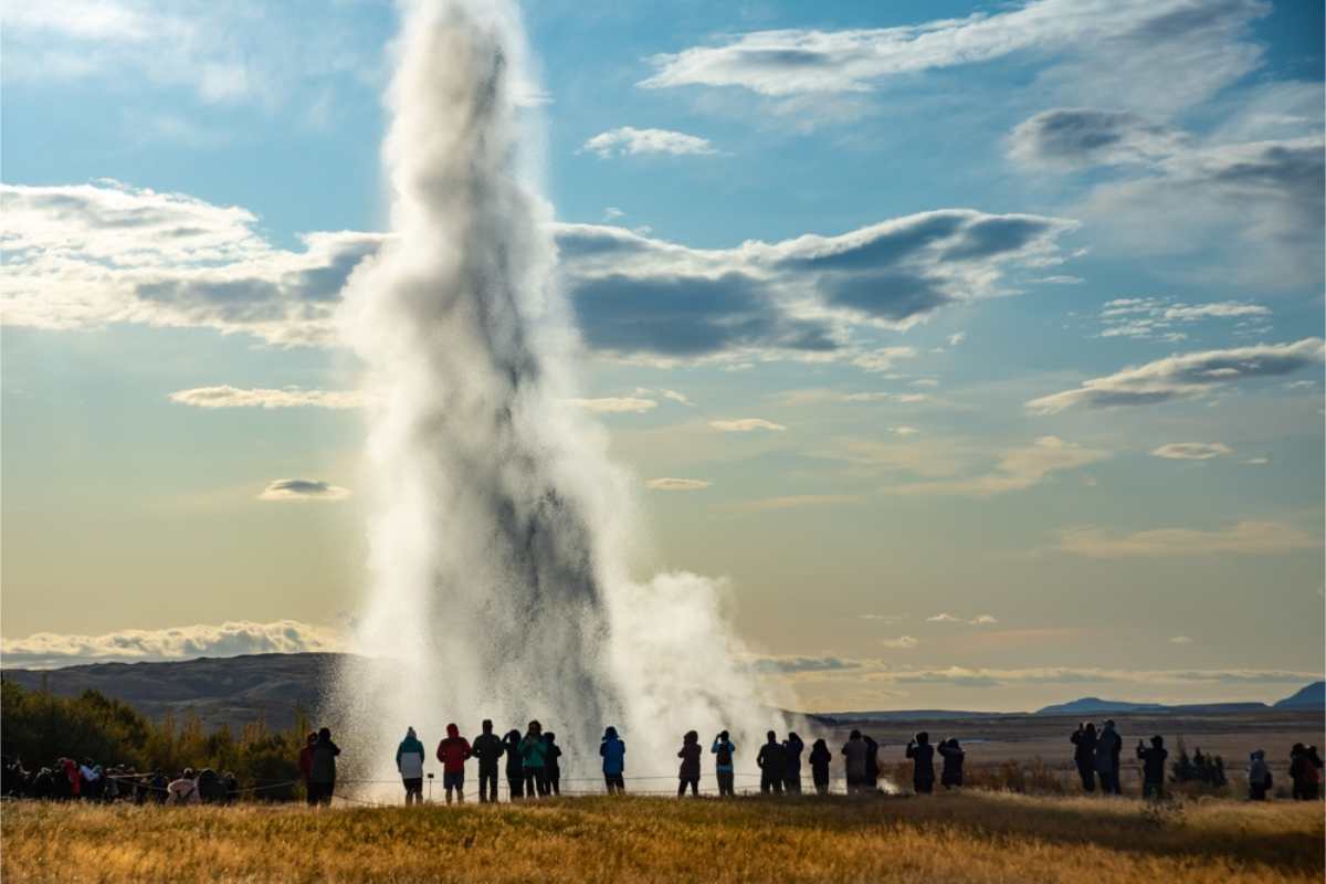 Turistas observan la erupción de un géiser, que lanza una enorme columna de agua y vapor al cielo bajo nubes dispersas.