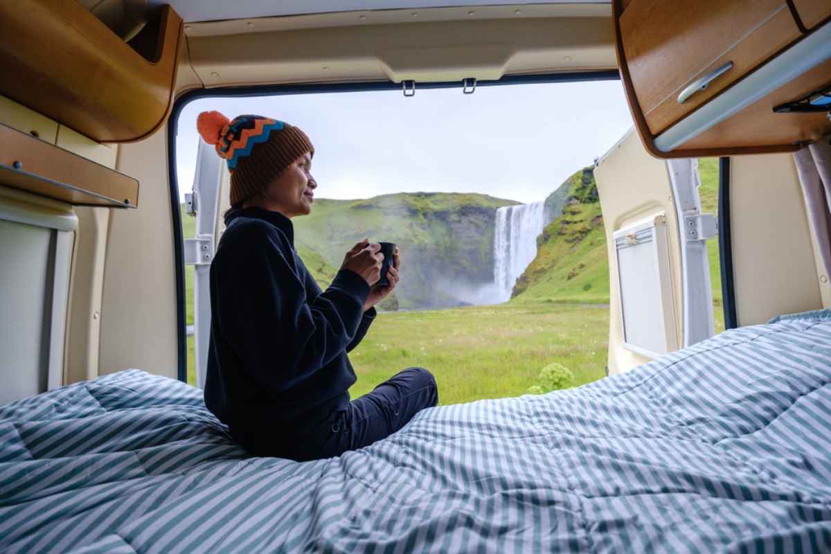 Viajero sentado dentro de una camper con una taza, mirando la cascada Skogafoss a trav&eacute;s de las puertas traseras abiertas