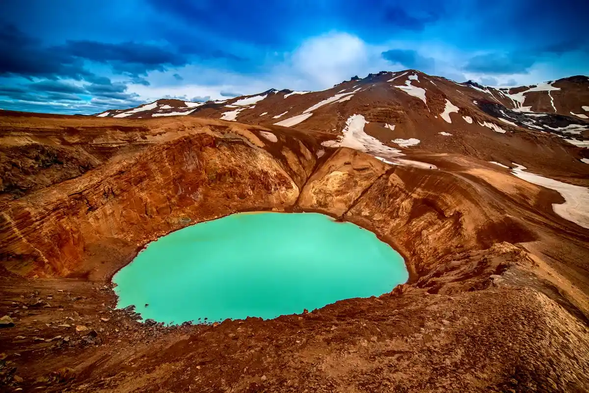 Un sereno lago de cr&aacute;ter turquesa rodeado de terrenos volc&aacute;nicos de color rojizo oscuro bajo un cielo azul profundo con rastros de nieve en los picos lejanos.