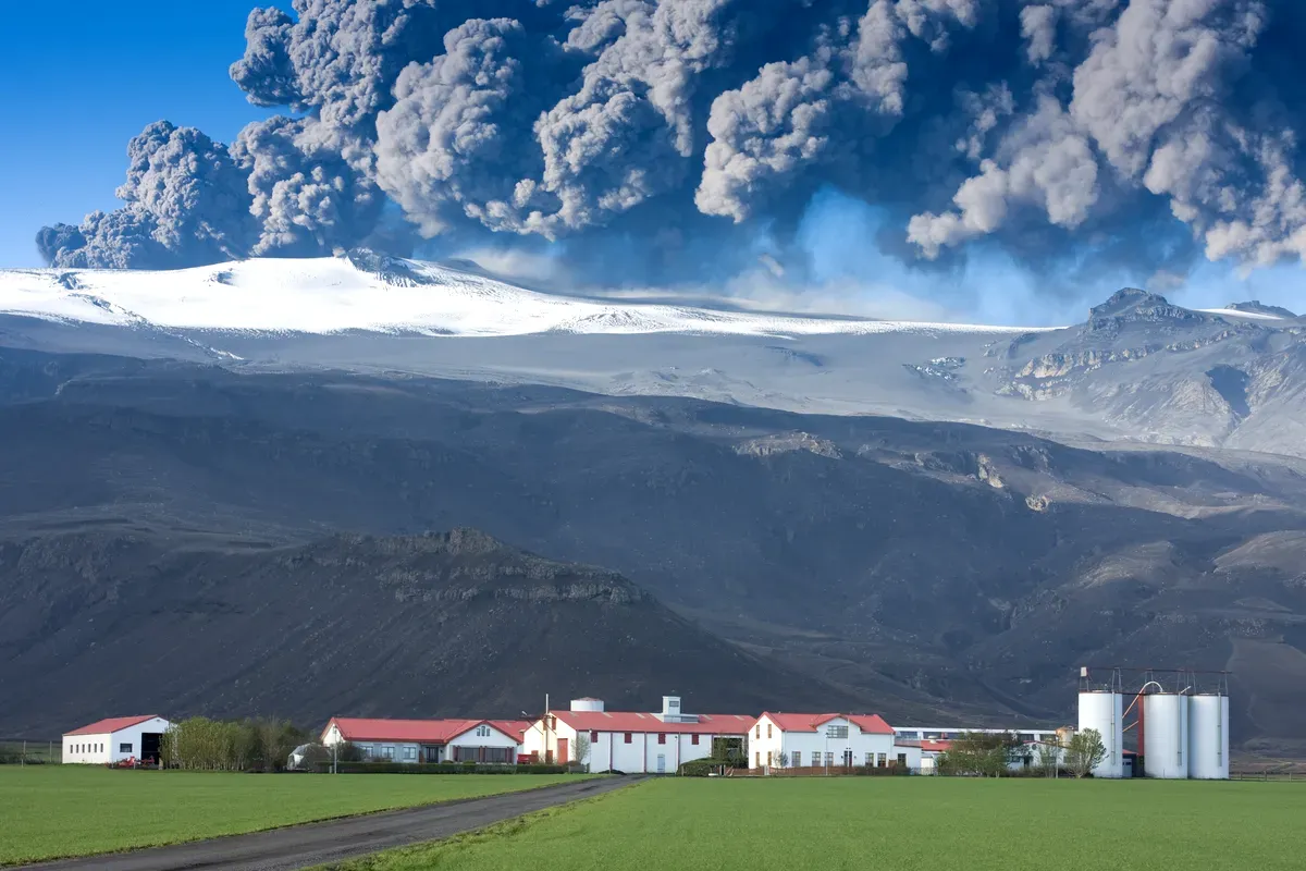 Una alta columna de ceniza y humo se eleva hacia el cielo desde el Volc&aacute;n Eyjafjallaj&ouml;kull, con una granja pac&iacute;fica y un campo verde en primer plano, contrastando con el volc&aacute;n en erupci&oacute;n.