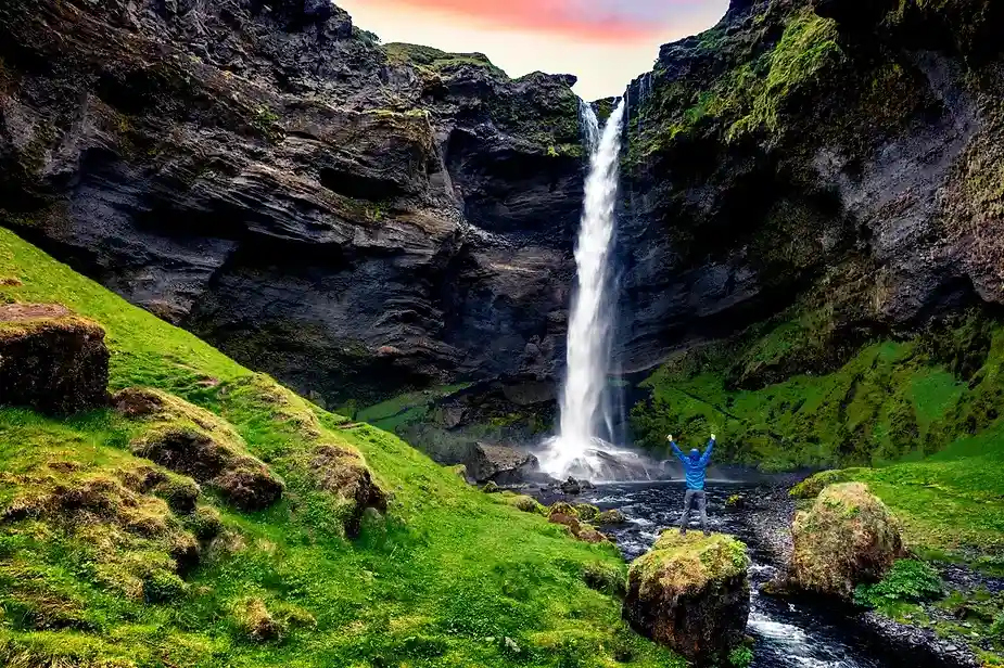 Senderista de pie con los brazos levantados frente a la cascada de Kvernufoss, rodeado de acantilados cubiertos de musgo y exuberantes paisajes verdes