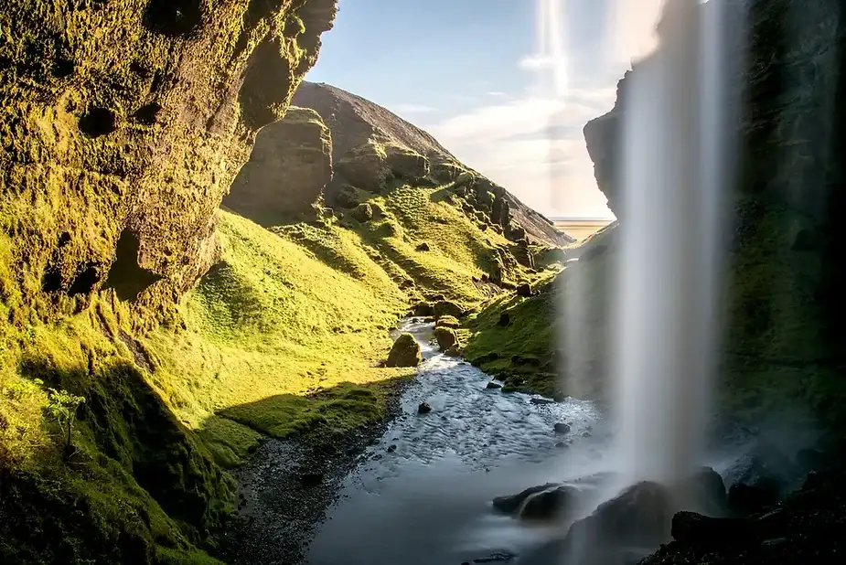 Luz del sol brillando a trav&eacute;s de la niebla de la catarata Kvernufoss en Islandia, con un arroyo tranquilo fluyendo por un valle verde y acantilados en el fondo.