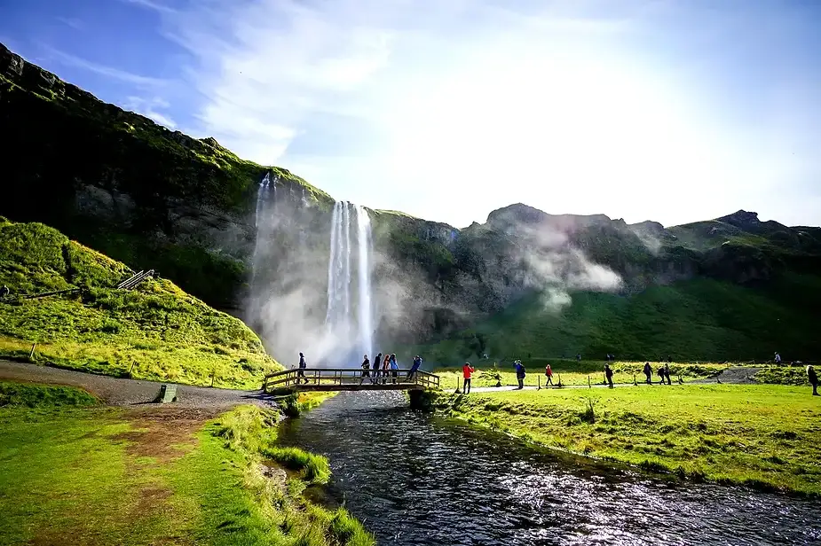 Turistas cruzando un puente de madera cerca de la cascada Seljalandsfoss en Islandia, con exuberantes paisajes verdes y un cielo azul brillante.