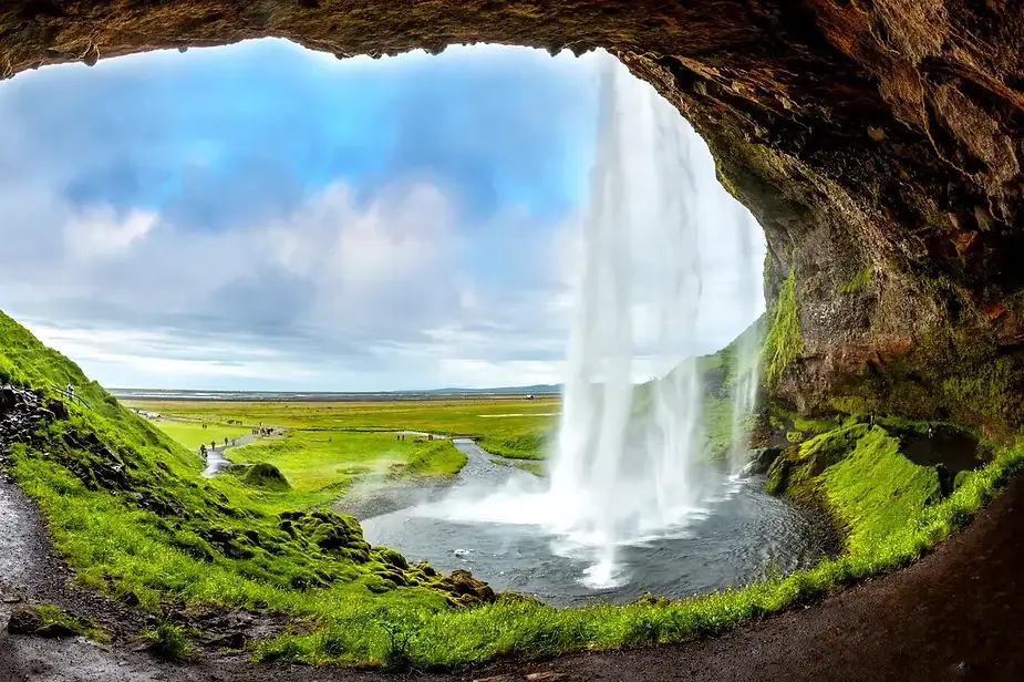 Vista desde detr&aacute;s de la cascada Seljalandsfoss en Islandia, mostrando el exuberante paisaje verde y un cielo azul nublado