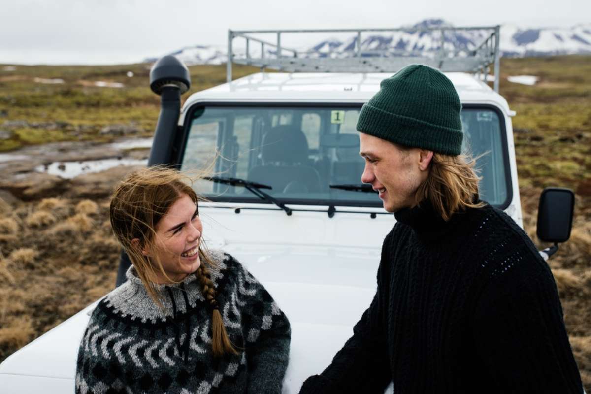 Deux voyageurs discutent &agrave; c&ocirc;t&eacute; d&rsquo;un 4x4 blanc dans les Hautes Terres islandaises, avec un sol couvert de mousse et des montagnes enneig&eacute;es en arri&egrave;re-plan.