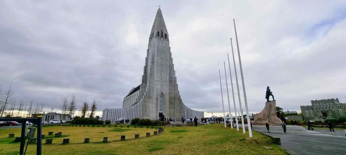 Église Hallgrímskirkja à Reykjavík Hallgrímskirkja à Reykjavik avec la statue de Leif Erikson et de hauts mâts; grande place et stationnement sur rue tout proche sous un ciel gris.